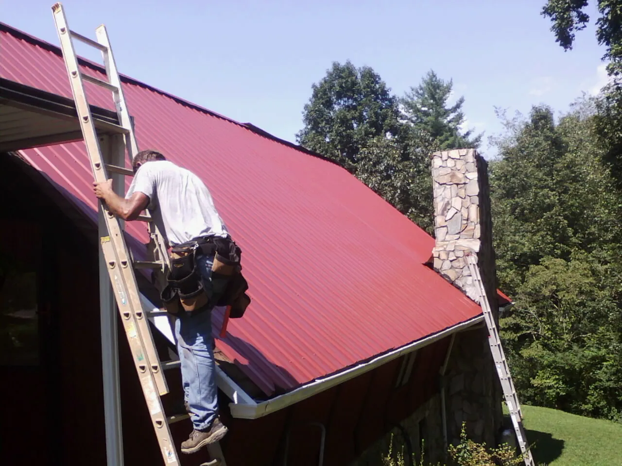 Roofer on a ladder beside a red metal roof and stone chimney — Soffit & Fascia Repair by Roofing Brothers in Lake Hughes